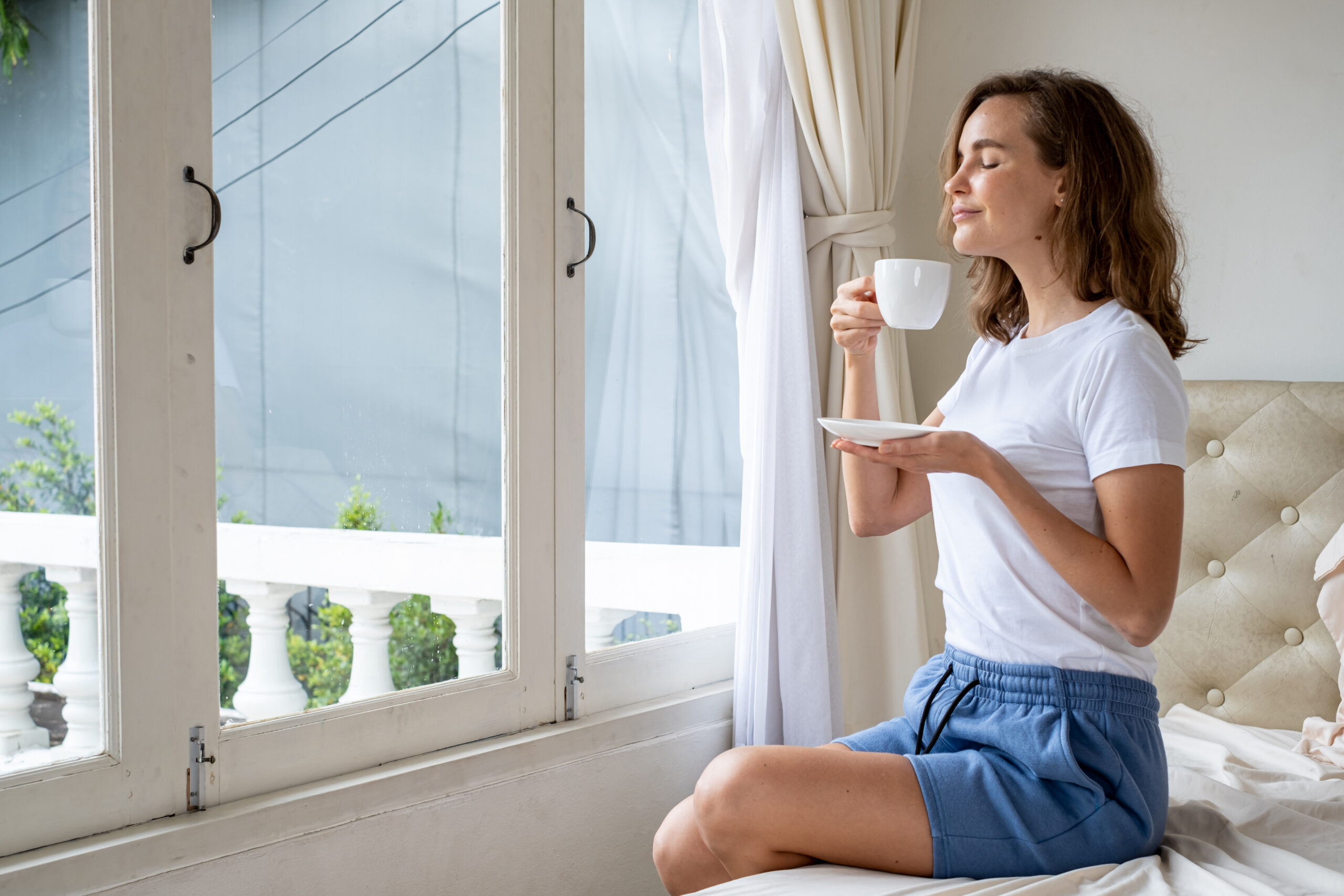 A woman is drinking coffee while sitting on the bed in her bedroom at home and relaxing in front of the windows.