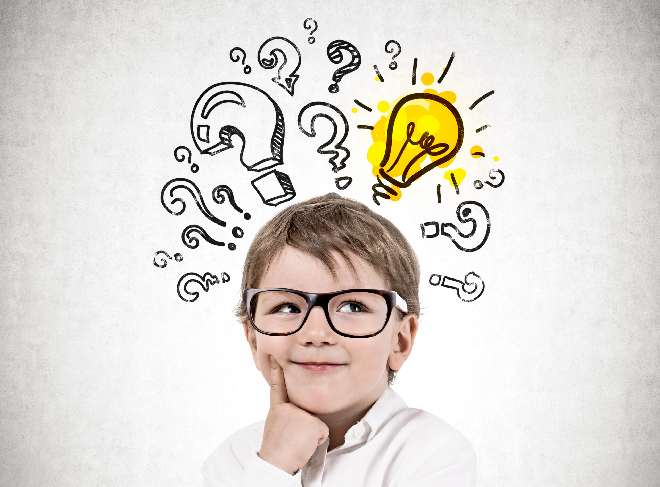 Adorable blond little boy wearing glasses and thinking standing near concrete wall with many question marks and lightbulb. Concept of idea