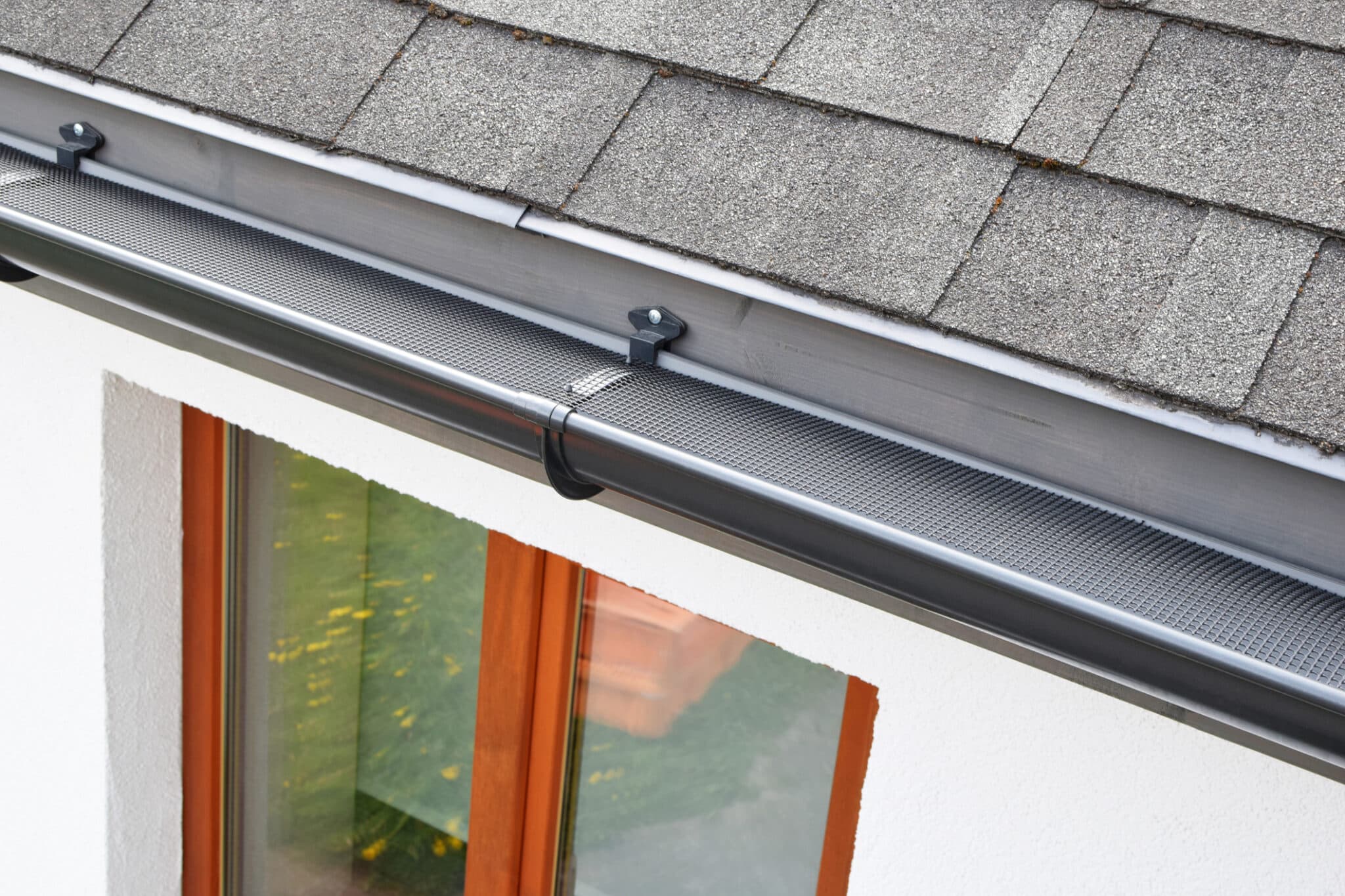 A gutter guard is installed over a new dark grey rain gutter on an asphalt shingle roof.